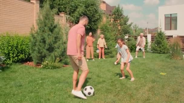 スローモーションフルショットの若いです大人男ドリブルで芝生の上で大きな世代の家族は晴れた夏の日に裏庭でサッカーをしています — ストック動画