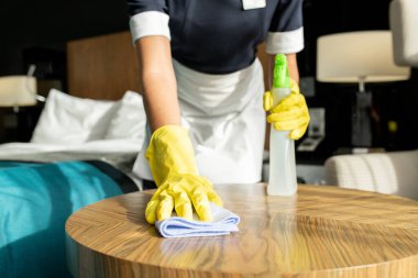 Çağdaş oda hizmetçisi üniformalı ve koruyucu eldivenli. Otel odasındaki ahşap masayı temizlerken deterjan ve deterjan kullanıyor.