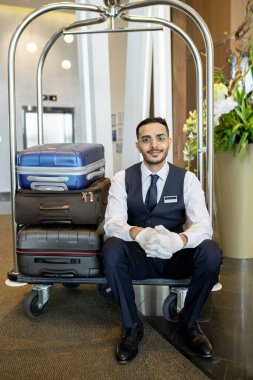 Üniformalı, eldivenli ve gözlüklü genç hamal modern otelin koridorunda, kameranın önünde misafirlerle el arabasında oturuyor.