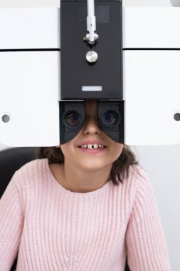 Modern kliniklerde gözleri kontrol edilirken göz hekimliği ekipmanlarının önünde oturan mutlu kız öğrencinin yüzü.