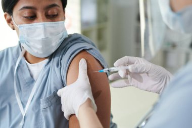 Çağdaş kliniklerin tıbbi muayenehanesinde aşı sırasında melez kadın meslektaşına iğne yapan eldivenli bir klinisyen.