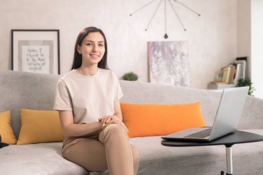 Küçük bir masanın önünde dizüstü bilgisayarla oturan ve oturma odasında dinlenirken dişlek bir gülümsemeyle sana bakan mutlu genç kadın.