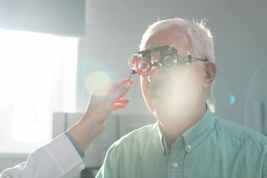 Kadın göz doktorunun önünde oturan kıdemli bir adam klinikteki optometrist deney çerçevesinde görme yeteneğini kontrol ediyor.
