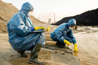Koruyucu tulumlar içinde çağdaş ekologlar zehirli ve kirlenmiş suyun özelliklerini incelerken, dizüstü bilgisayarlı bir kadın yeni veri girişi yapıyor.