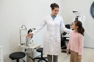 Whitecoat 'taki genç ve mutlu esmer göz doktoru küçük kıza yeni test ekipmanları gösteriyor ve kliniklerde görme yeteneğini kontrol etmesi için onu davet ediyor.