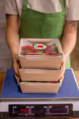 Genç, dikey tarım işçilerinin elleri taze çilek dolu konteynırları paketledikten sonra onları teraziye koyarken tutuyorlar.