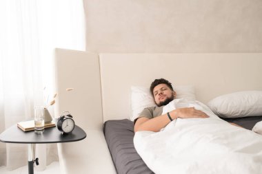 Tişörtlü genç sakallı adam, büyük, rahat, çift kişilik bir yatakta, çalar saati, bir bardak suyu, vazosu ve kitabı olan küçük bir masanın önünde uyuyor.