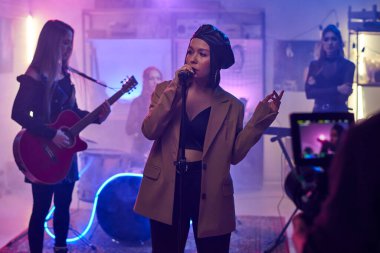 Kadın şarkıcı mikrofonla şarkı söylüyor ve grubuyla birlikte şarkı söylüyor ve konserlerini videoda kaydediyor.