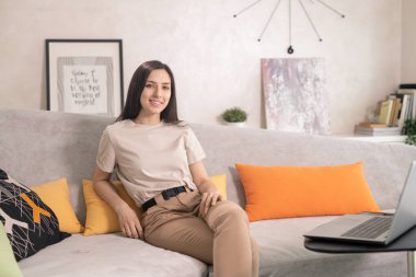 Koyu renk uzun saçlı genç bir bayan oturma odasında küçük bir masanın önünde oturuyor ve sana bakıyor.