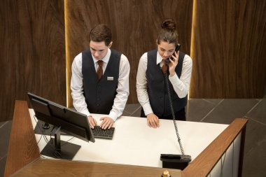 İki genç otel resepsiyonistinden biri tezgahın yanında durup bilgisayar ekranına bakarken iş arkadaşı da yakınlardaki telefona cevap veriyor.