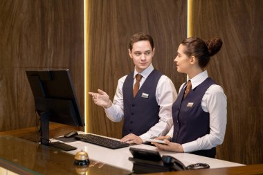 Tezgahta duran iki genç otel resepsiyonistinden biri dokunmatik bilgisayarlı meslektaşına bakıyor ve bilgisayar ekranını işaret ediyor.