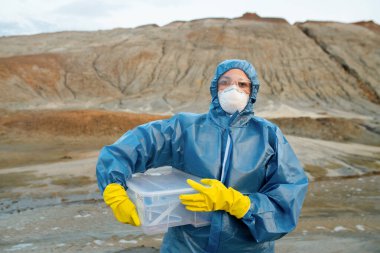 Koruyucu tulumlu, solunum cihazlı, lastik eldivenli ve gözlüklü, içinde su ve toprak örnekleri olan plastik kap tutan genç bir araştırmacı.
