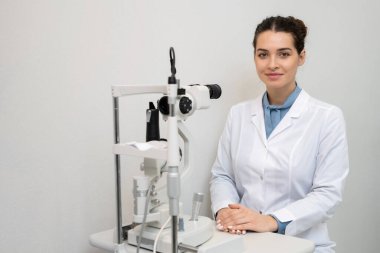 Whitecoat 'ta gülümseyen genç bayan göz doktoru kliniklerde tıbbi ekipmanların önünde oturmuş ve hastaları bekliyor.