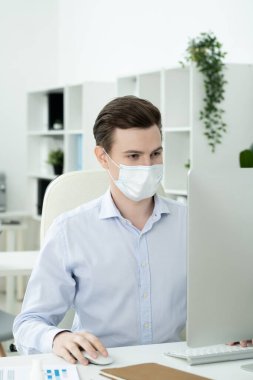 Yüzünde koruyucu maske olan genç ciddi erkek tasarımcı ofis ortamında masa başı bilgisayar ekranına bakıyor.