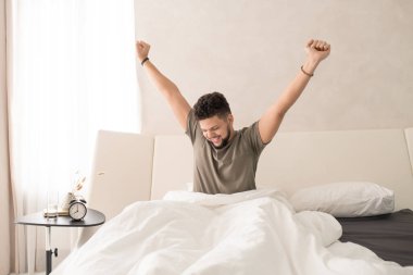 Sabahları beyaz battaniyenin altında büyük ve rahat bir çift kişilik yatakta otururken mutlu genç adam kollarını yukarı doğru açıyor.