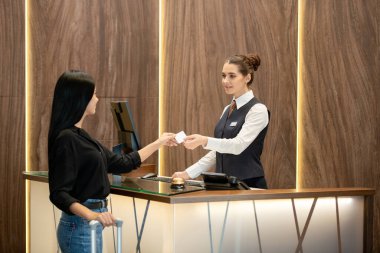 Üniformalı genç esmer resepsiyonist, iş yerinin yanında duruyor ve otel lobisindeki resepsiyondaki misafirlerden birine plastik kart uzatıyordu.
