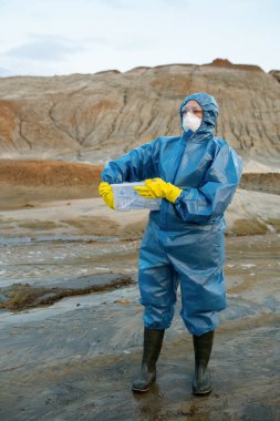 Genç kadın bilim adamı, tepeler ve nehirlere karşı kirli bölgede dururken su ve toprak örnekleriyle plastik kap tutuyor.