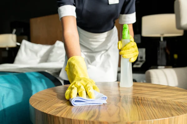ホテルの部屋で木製のテーブルを掃除しながら ダスターと洗剤を使用した制服と保護手袋の若い現代的な部屋メイド — ストック写真