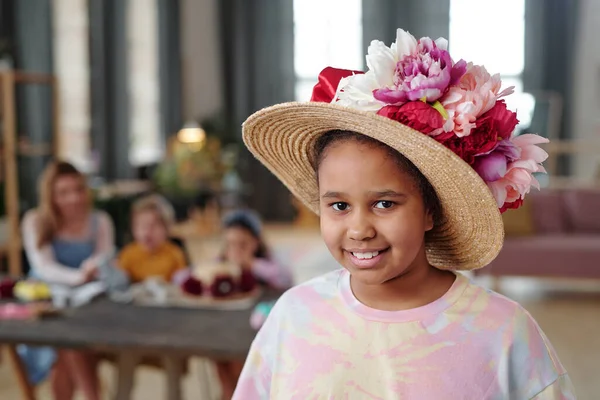 テーブルによって彼女の友人に対して立って白い ピンクと深紅の色の大きな手作りの花で飾られた帽子のかなり小さな混合レースの女の子 — ストック写真