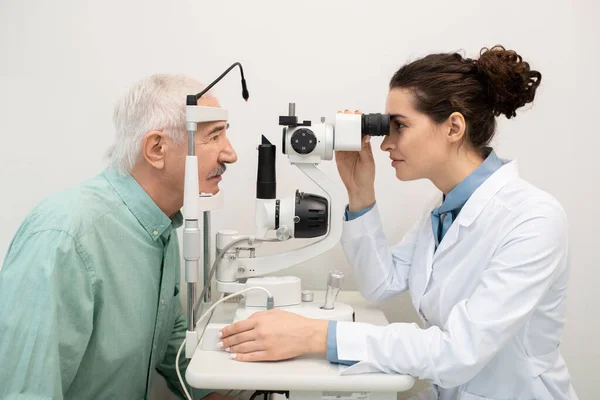 若いですブルネット女性眼科医によって座っています医療機器でクリニック中にテストしながら視力のシニア男性患者で彼女の前 — ストック写真