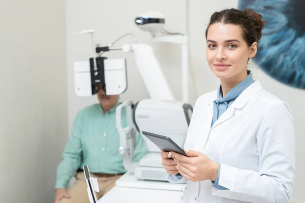 彼の視力をチェックしている患者の背景にタブレットを使用している間 あなたを見てホワイトコートの幸せな若い女性眼科医 — ストック写真