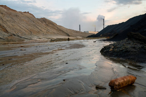 View of dirty road with mud and big puddles or river surrounded by polluted hills and industrial factories exhausting toxic fumes