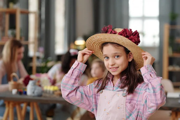 戴着帽子 装饰着大大的手工红花的漂亮小女孩站在桌旁笑着看着你和她的朋友们在一起 — 图库照片