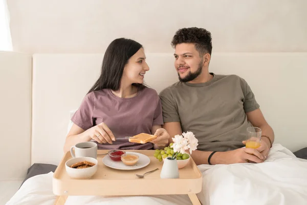 幸せな若い愛情のカップルでTシャツのベッドに座って朝食中に話す一方 女性はトーストにピーナッツバターを広げて — ストック写真