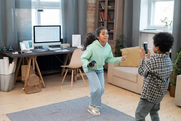 快乐的混血女孩在客厅中央的地板上开心地跳舞 而她的哥哥则用智能手机拍摄视频 — 图库照片