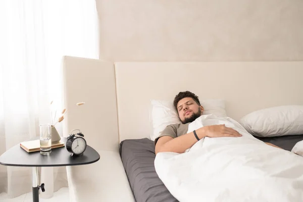 Young bearded man in t-shirt sleeping in large comfortable double bed in front of small table with alarm clock, glass of water, vase and book