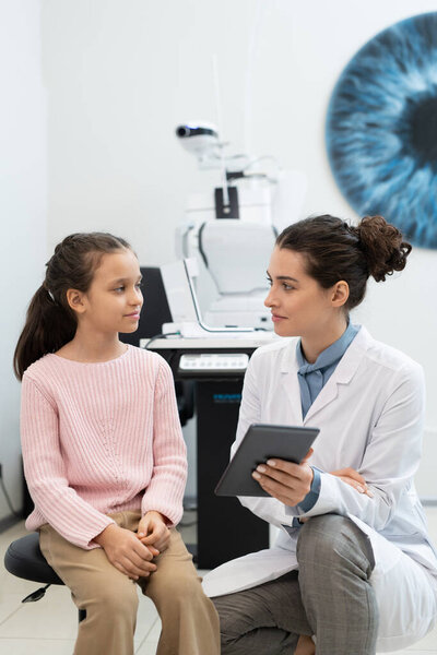 Young female ophthalmologist in whitecoat using digital tablet while showing schoolgirl results of check-up of her eyesight in clinics