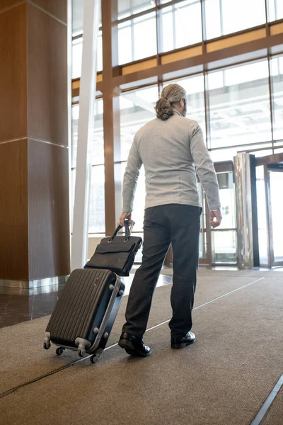 現代的なホテルの出口に向かって移動しながら 彼自身の後ろにスーツケースを引っ張っカジュアルで成熟したビジネスマンの背面ビュー — ストック写真