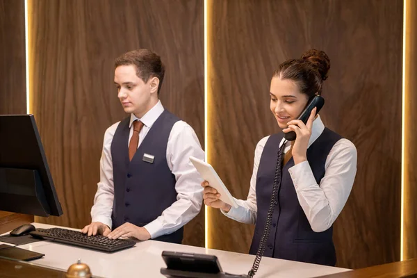 Tezgahın yanında duran iki genç otel resepsiyonistinden biri dokunmatik ekranlara bakıyor ve telefonda meslektaşlarına karşı danışmanlık yapıyor.