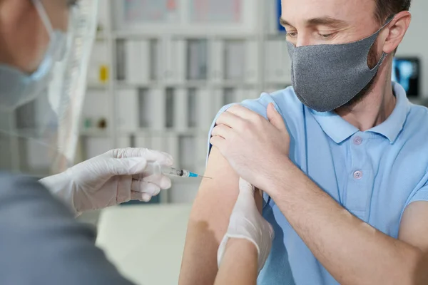 若い男は彼のシャツの袖を保持しながら 保護作業服で看護師は彼がクリニックでCovidに対する予防接種中に注射を作る — ストック写真