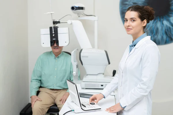 快乐的年轻女性眼科医生站在视力测试设备前 转动旋钮 看着你与老年男性患者对视 — 图库照片