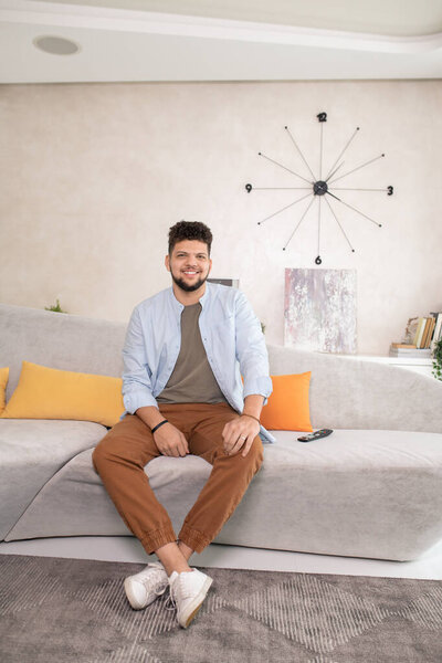 Happy young bearded male in casualwear looking at you with toothy smile while sitting on couch in front of camera in living-room