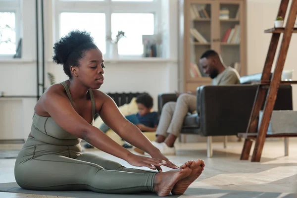 在与丈夫和小儿子的体育锻炼中 穿着运动服的年轻非洲妇女坐在客厅地板上的垫子上 — 图库照片
