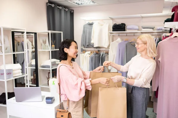 アジアの若い顧客は店で服を購入し セールスマンは彼女のショッピングバッグを笑顔で与えます — ストック写真