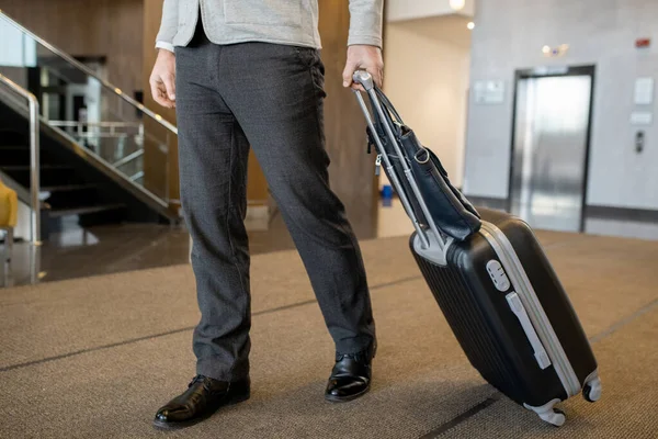 Low section of modern businessman in casualwear pulling suitcase behind himself while moving along hall of large and luxurious hotel