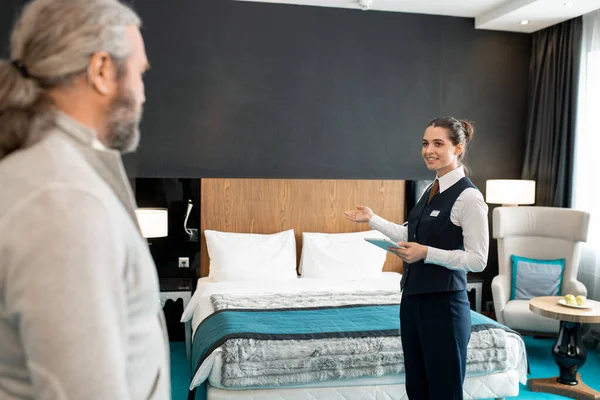 Young smiling hotel worker in uniform holding digital tablet while showing mature businessman his room and wishing him good rest