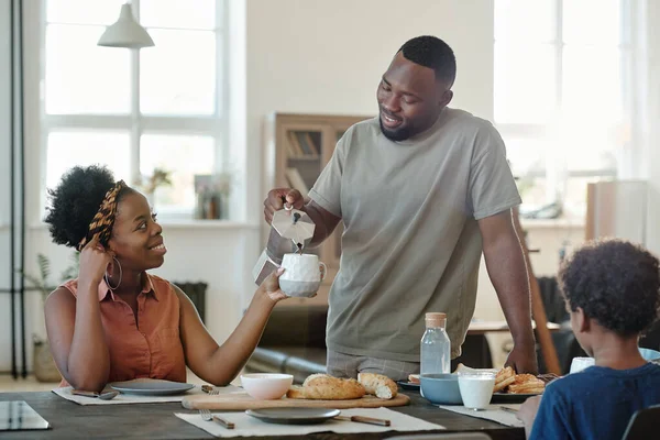 当天早晨 年轻的非洲男子站在厨房桌旁 在家人的早餐中 将茶或咖啡倒入妻子的杯子中 — 图库照片