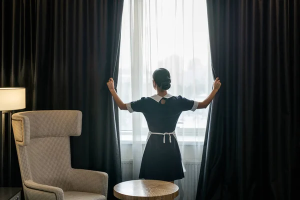 ホテルの部屋の大きな窓にかかっている均一な開口部の暗いカーテンで若い部屋のメイドのバックビューそれをきれいにする前に — ストック写真