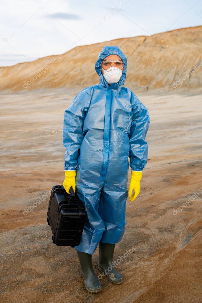 Young female researcher in protective workwear holding case with samples while standing on polluted soil in abandoned area against hill and sky