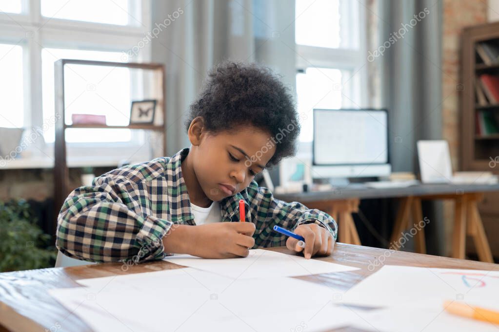 Diligent schoolboy in casualwear drawing with highlighters on paper by table in living-room while enjoying leisure after doing homework