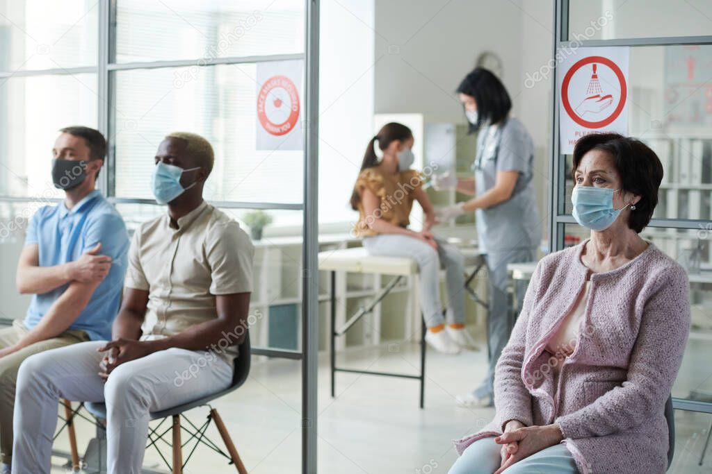 Mature woman and other patients in protective masks sitting on chairs in clinics and waiting for their turn to have vaccination against covid