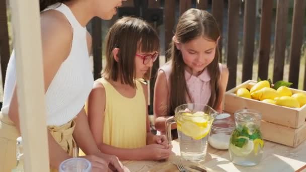 夏の日に屋外で新鮮なレモネードを作る2人のかなり10歳の姉妹の中ショット 水とレモンとガラスのデカンタに砂糖を追加し お母さん 彼らを助ける — ストック動画