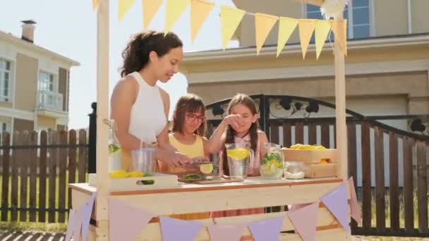 夏日炎热的时候 站在户外市场的木制灯台上 用柠檬和薄荷做了一杯清爽的柠檬水 拍下了一位快乐的混血女子和两个漂亮的小女儿的照片 — 图库视频影像