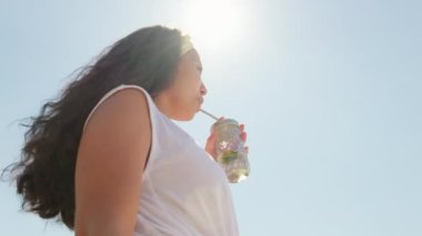 Açık havada duran uzun kıvırcık saçlı ve yaz tatilinin tadını çıkaran limonatasını yudumlayan çekici genç bir kadının orta açı çekimi.