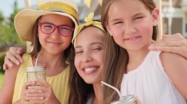 Çekici genç bir kadının yakın plan portresi ve 10 yaşında iki güzel kız sıcak yaz gününde dışarıda kameraya gülümsüyor, limonata içen kızlar.