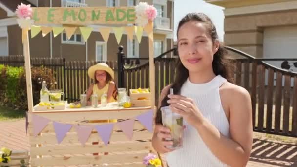 在炎炎夏日里 一位漂亮的混血女子在户外喝着新鲜柠檬水 而一位10岁的漂亮姑娘则站在后台卖清爽的饮料 — 图库视频影像